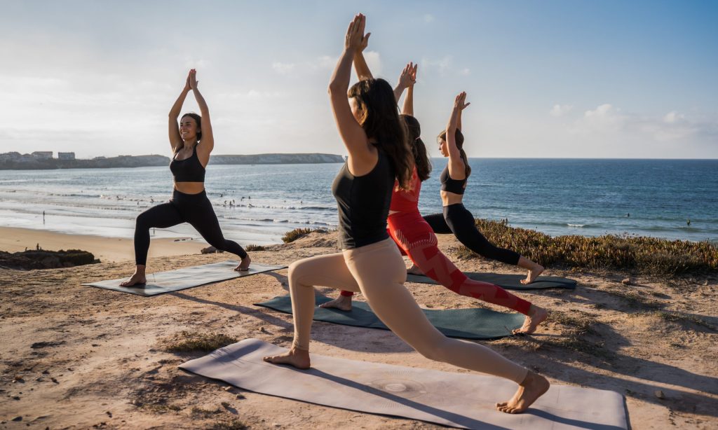 Group of young woman fitness instructor in sportswear stretching at the beach on a yoga mat with her group of students. Female fitness yoga routine concept