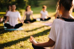 Group of young sporty woman practicing yoga lesson with instructor, sitting in lotus pose. Focus of camera at teacher rear view. Photo from back of instructor. Group of meditating people in background
