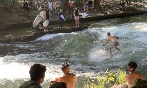 Surfer riding a standing river wave with swimmers and spectators watching from the bank