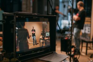 Film camera monitor in the foreground showing a scene, with the set and crew visible behind it