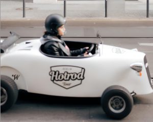 Driver in a helmet at the wheel of a small white Hot Rod car with the Hotrod logo on the door, seen from the side on a stree