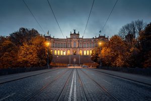 Illuminated neoclassical building with a columned facade at dusk, tram tracks and autumn trees in the foreground