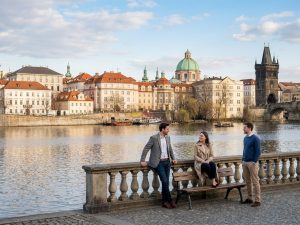 Colleagues talking beside the Vltava with the Old Town skyline and a Charles Bridge tower behind
