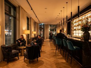 Hotel bar in Prague with a long counter, teal bar stools and armchairs beside tall windows