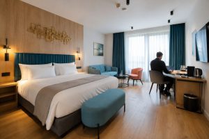 Hotel bedroom in Budapest with a white double bed, teal cushions and a guest working at the desk by the window