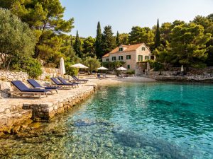 Stone boutique property among pine trees, with sun loungers and parasols on a terrace beside a turquoise cove