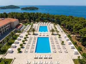 Long rectangular hotel pool lined with sun loungers and palm trees, beside a terracotta-roofed wing with the sea beyond