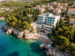 Modern white hotel terraced into a pine-covered headland above a turquoise Adriatic cove, with a small jetty at the shoreline