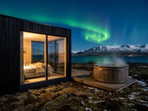 Dark-clad boutique cabin lit from within with an outdoor hot tub and Northern Lights overhead
