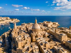 Elevated view over honey-coloured limestone rooftops in Malta with a church dome and the sea beyond