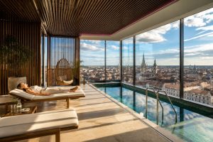 Indoor rooftop pool with sun loungers, a hanging rattan chair and floor-to-ceiling windows over the city rooftops