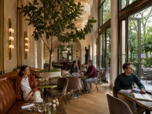 Diners at round tables in a light-filled restaurant with a large indoor tree and tall arched windows
