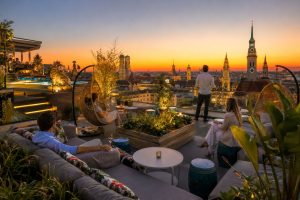 Guests on rooftop terrace sofas at sunset with the Frauenkirche and old town skyline behind, Munich