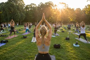 Large outdoor yoga class on mats in a park at sunrise, participant in the foreground with arms raised overhead
