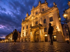 Illuminated baroque façade on a cobbled Prague square at dusk with people walking past