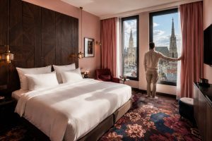 Hotel bedroom with a white bed, dark panelled headboard wall and rose curtains framing a city-view window