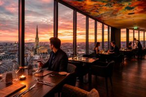 Guests dining beside floor-to-ceiling windows in an upper-floor restaurant overlooking the city skyline at sunset